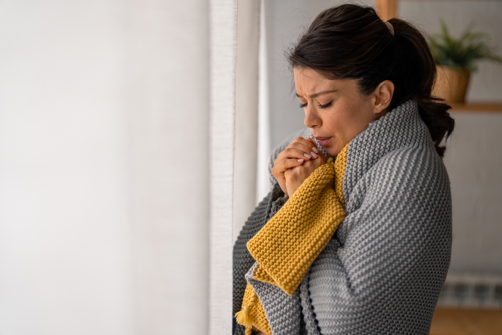 A woman wrapped in a blanket and blowing on her clutched fists because it's cold in her house