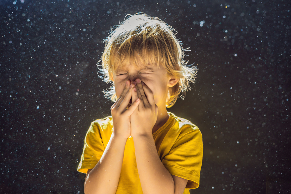 A little boy sneezing into his hands as dust floats in the air behind him