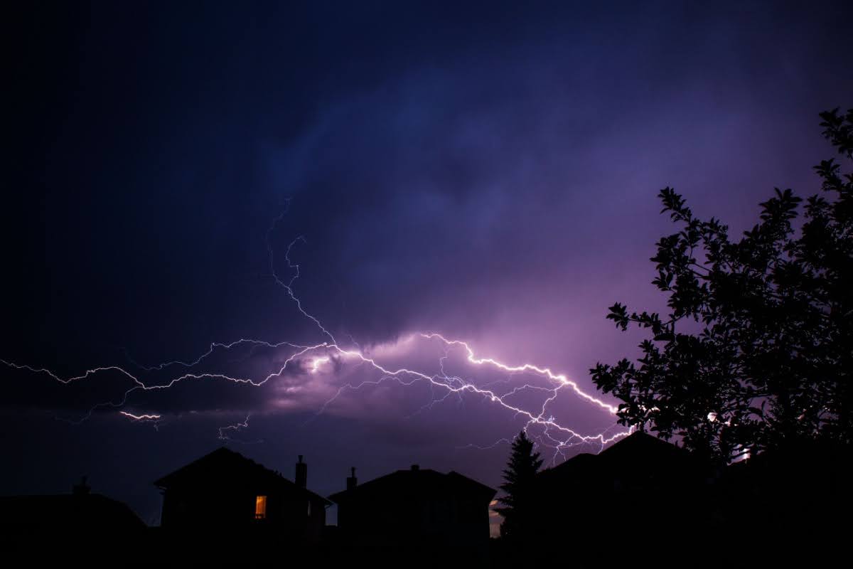 A lightning strike lighting up a lavender/purple sky above the silhouettes of homes