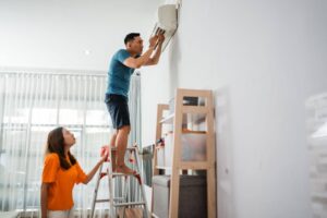 Husband standing on a ladder trying to troubleshoot a ductless mini split unit on the wall while his wife stands at the foot of the ladder