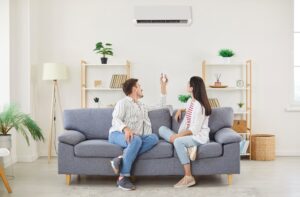 Couple sitting on a gray couch and twisting around to look at their AC unit mounted on the wall, which isn't turning on