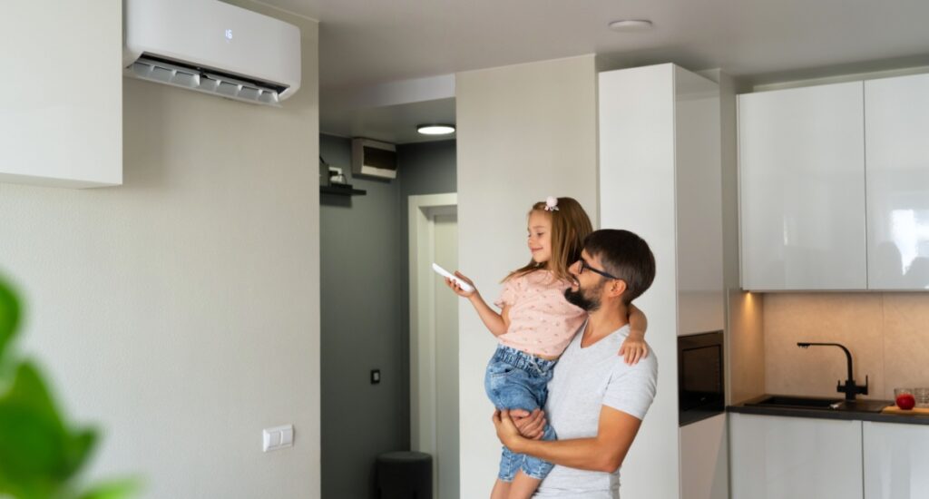A dad holding his young daughter has she uses a remote to turn on a new AC unit on the wall