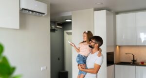 A dad holding his young daughter has she uses a remote to turn on a new AC unit on the wall