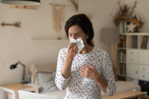 woman sitting on her bed and blowing her nose into a tissue because of the indoor allergens