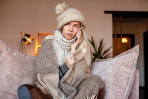 a cold woman wrapped in a blanket and wearing a wool hat while sitting on the couch and calling a heating technician